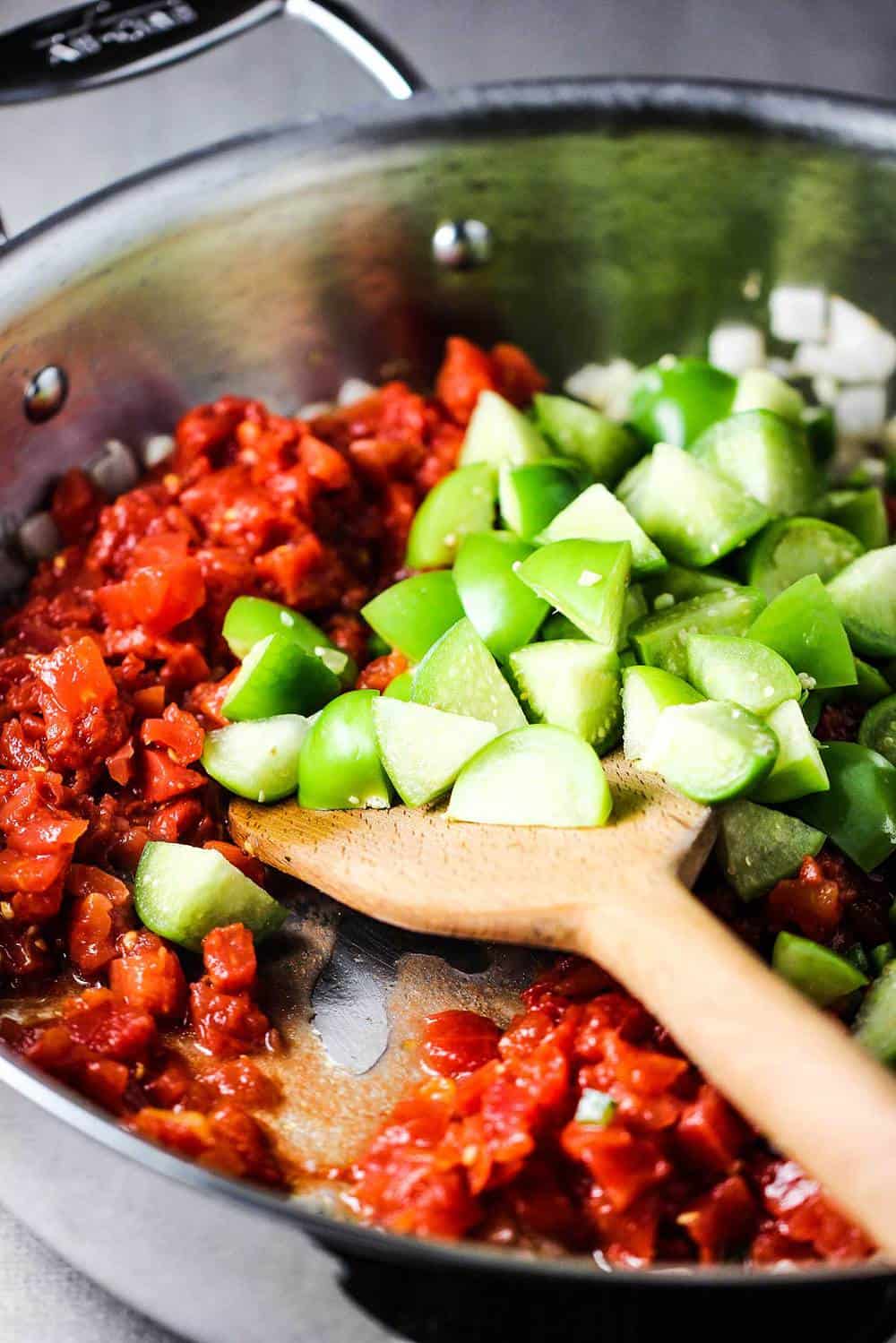 Chopped tomatillos, jalapeño and tomatoes in a skillet with a wooden spoon