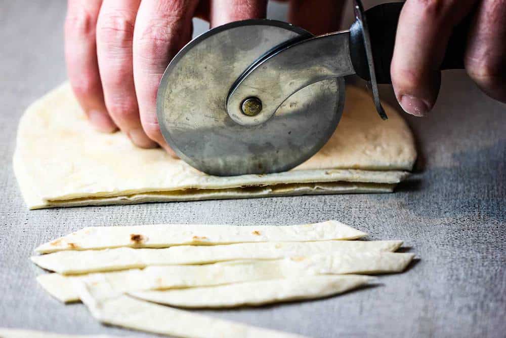 A hand using a pizza cutter to cut tortillas strips for white chili