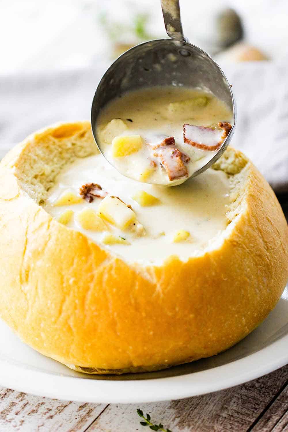 A ladle pouring New England Clam Chowder into a sourdough bread bowl on a white plate.