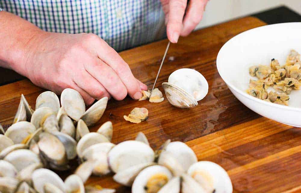 Two hands chopping clam meat on a cutting board next to opened clams.