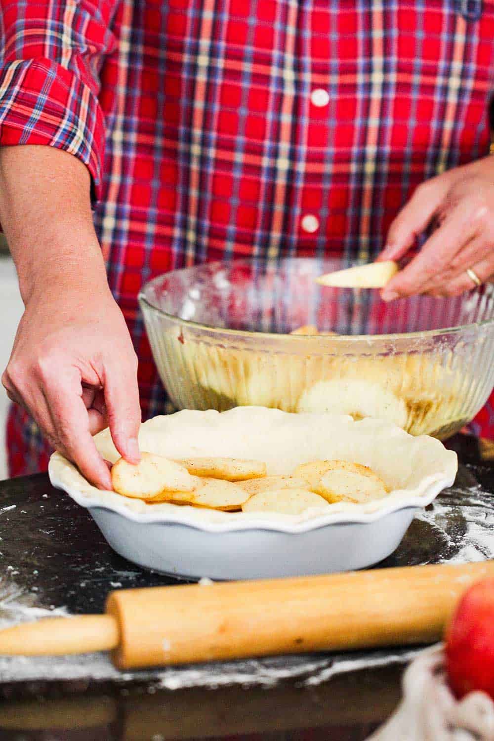A hand layering spiced apple slices into a pie dish lined with homemade pie dough. 