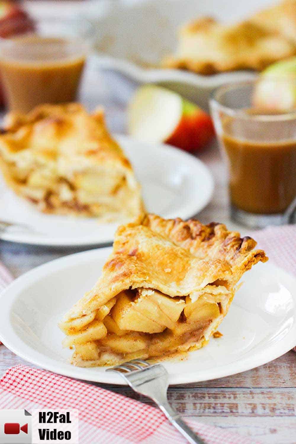 A slice of homemade apple pie on a white plate with a fork and another plate of pie behind it. 