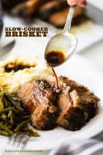 A spoon drizzling au jus sauce on slices of slow cooker brisket on a white plate.