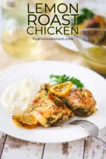 A white plate filled with two roasted chicken pieces, a lemon slice, and mashed potatoes next a glass of white wine.