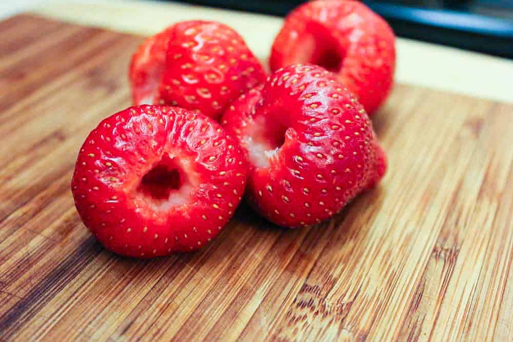 Four fresh strawberries that have been hulled and sitting on a wooden cutting board.