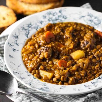 A white bowl of rustic lentil soup with slices of toasted bread stacked next to it.