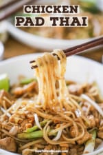 A pair of chopsticks lifting rice noodles from a bowl filled with chicken pad Thai.