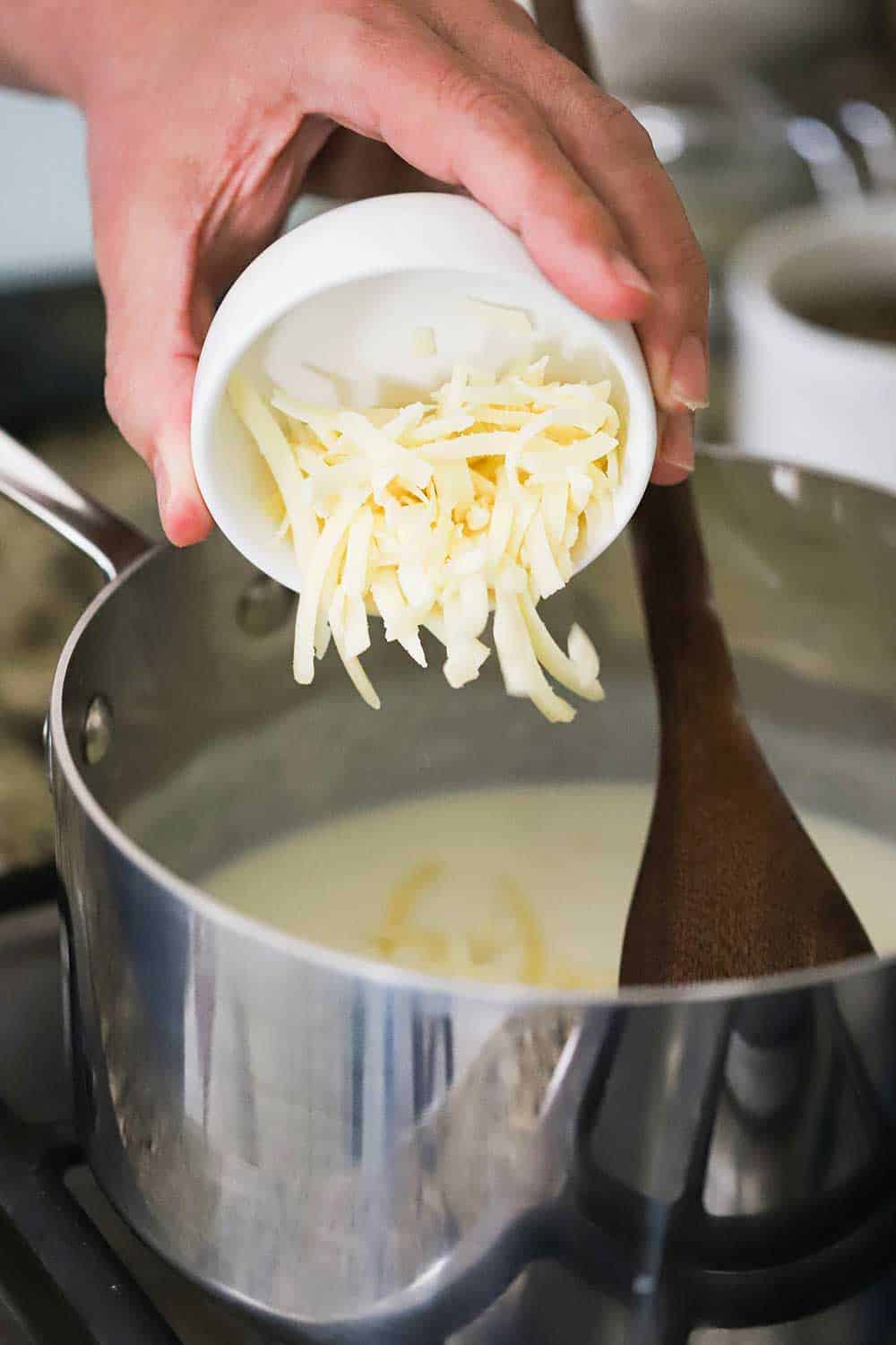 A hand transfer shredded Gruyere cheese from a small white bowl into a sauce pan filled with a béchamel sauce. 