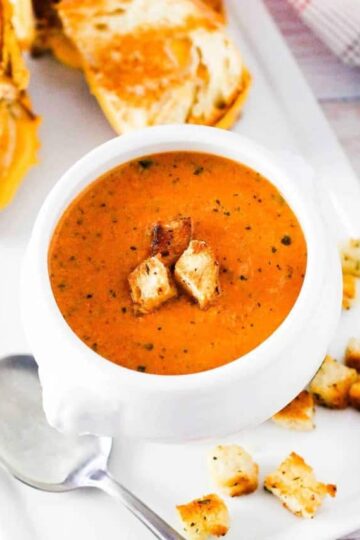 A white soup filled with roasted tomato basil soup next to sliced grilled cheese sandwiches.
