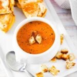 A white soup filled with roasted tomato basil soup next to sliced grilled cheese sandwiches.