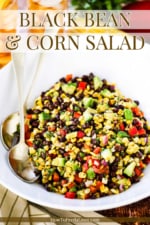 A large white serving bowl filled with black bean and corn salad with two gold serving spoons tucked in beside the salad.