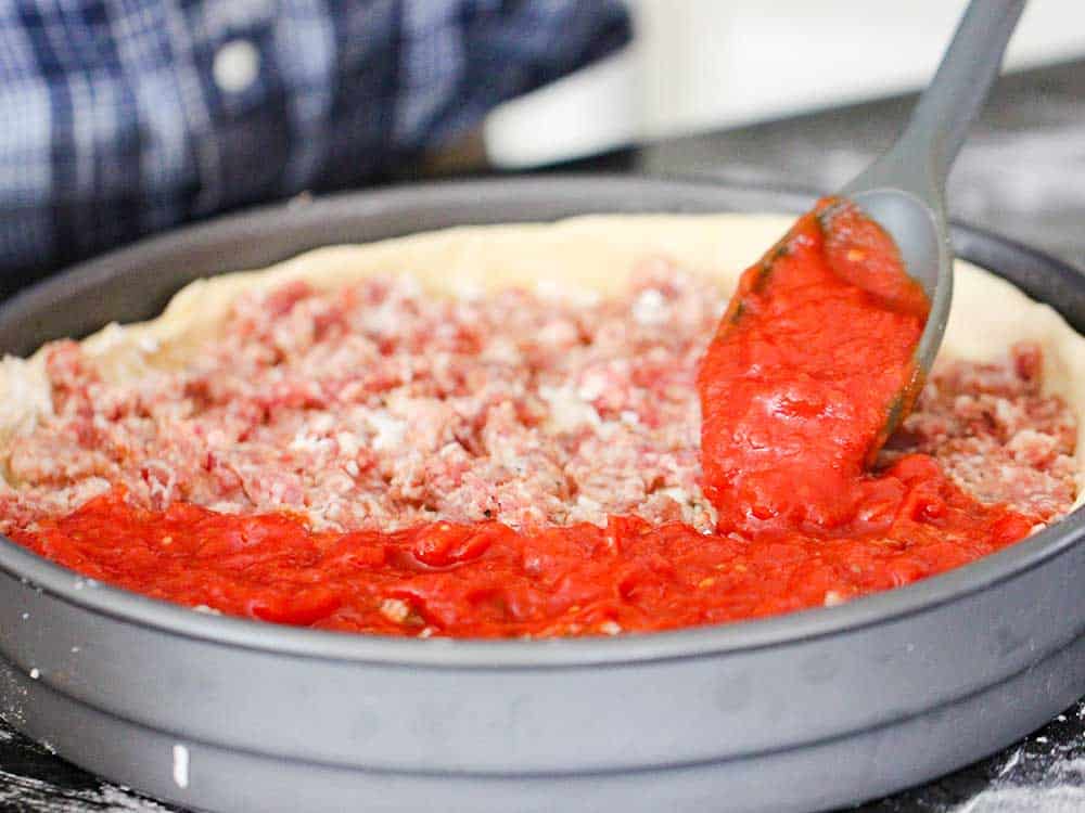 A spoon ladling pizza sauce over sausage in a deep dish pizza pan. 