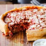 A Chicago-style deep dish pizza with a slice missing sitting on a wooden cutting board.