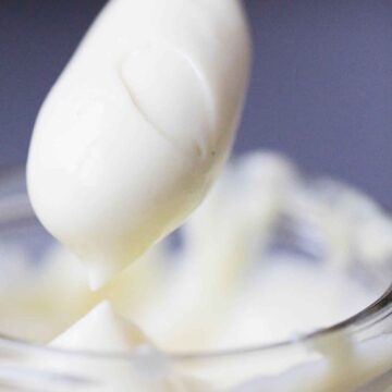 A spoon lifted from a clear container completely covered with homemade mayonnaise.