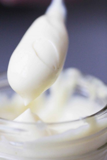 A spoon lifted from a clear container completely covered with homemade mayonnaise. 