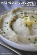 A circular white bowl filled with mashed cauliflower with roasted garlic topped with a pad of melting butter and snipped chives.