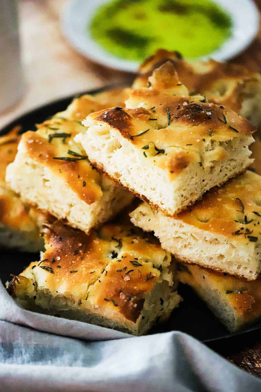 A black plate with a stack of square pieces of focaccia bread with rosemary and olive oil on it. 