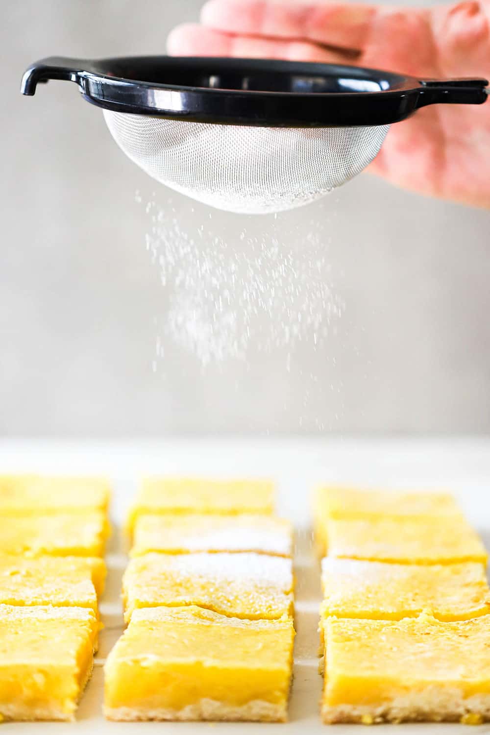 A hand using a small sieve to sprinkle powdered sugar over the tops of cut lemon bars. 