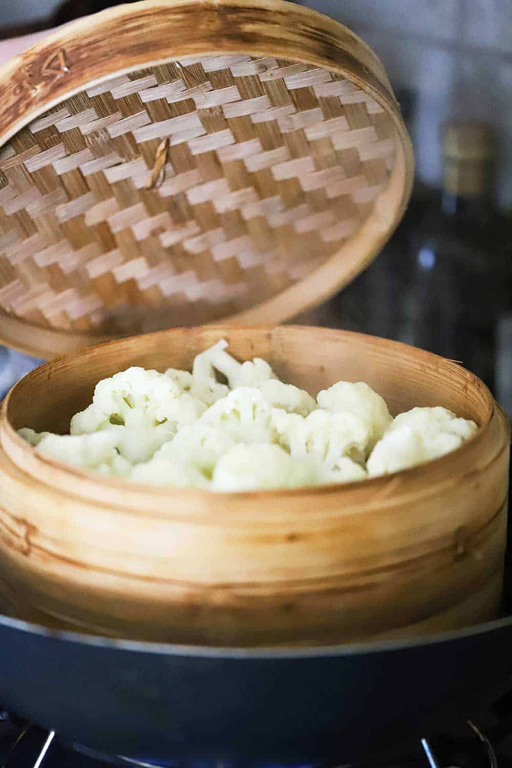 A lid of a bamboo steamer being lifted up to reveal cauliflower florets that have been steamed. 