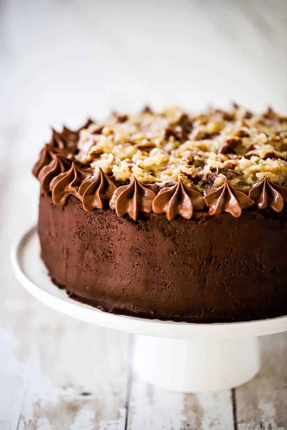 An unsliced German chocolate cake sitting on a white cake stand. 