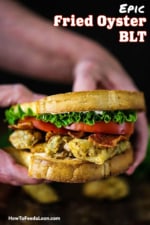 A person holding a Fried Oyster BLT consisting of two toasted slices of bread with crunchy oysters, crispy bacon, sliced tomatoes, and lettuce.
