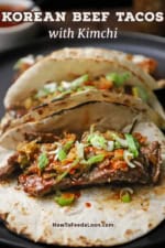 A straight-on view of three Korean beef tacos with kimchi on lightly charred flour tortillas all resting on a black dinner plate.