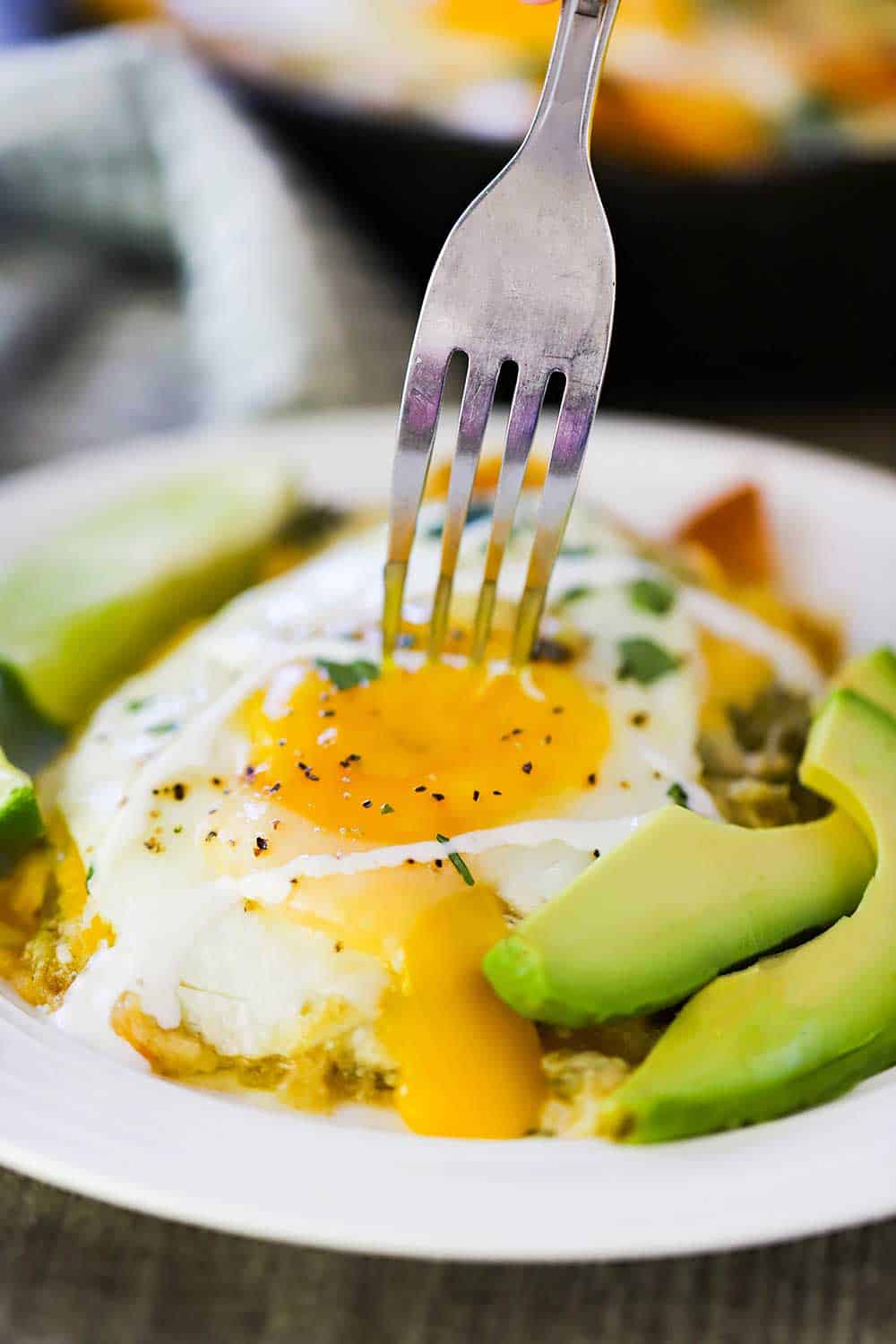 A fork piercing a sunnyside up fried egg onto of a plate of chilaquiles. 