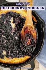 An overhead view of a cast-iron skillet containing blueberry cobbler with a portion of the cobbler missing and a wooden spoon inserted into the side.