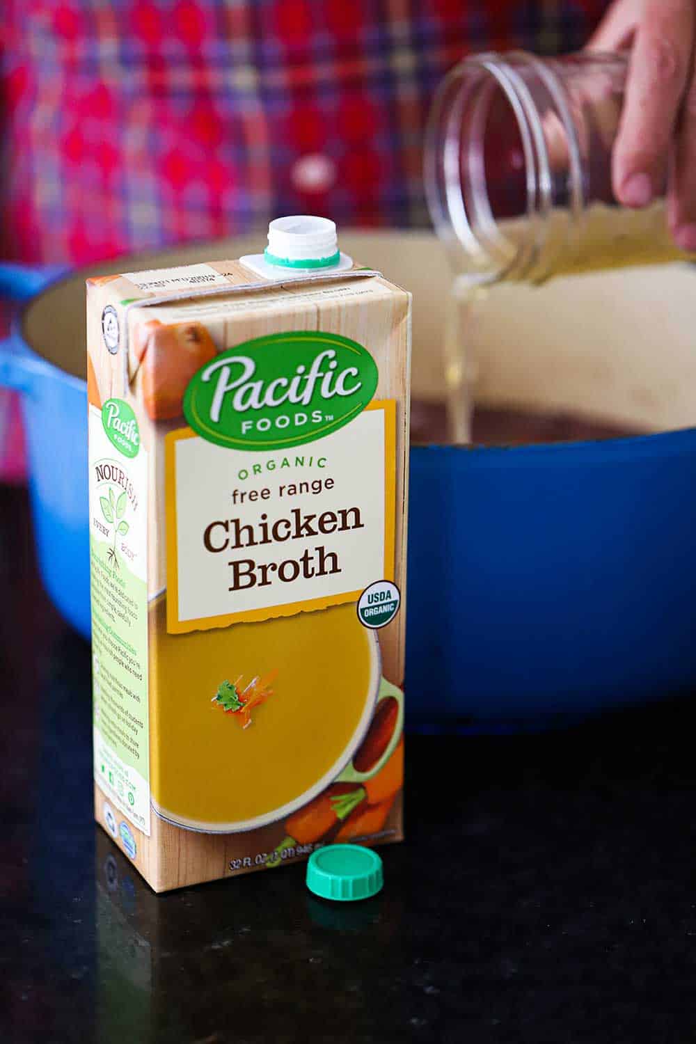A box of Pacific Food Chicken Broth in front of a blue Dutch oven with a hand pouring broth from a jar into the pot. 
