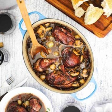 An overhead view of a large oval-shaped Dutch oven filled with Coq au Vin with a wooden spoon in and next to a cutting board topped with bread and next to a glass of red wine.