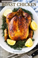 A roast chicken sitting on an oval platter with sliced lemons and herbs surrounding the bird.