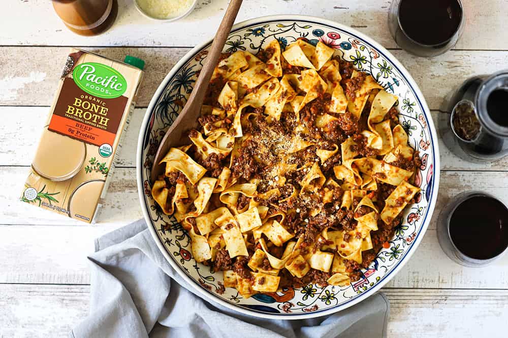 A large Italian pasta bowl filled with Pappardelle Bolognese sauce sitting next to a carafe and 2 glasses of red wine, and a box of Pacific Foods beef broth. 