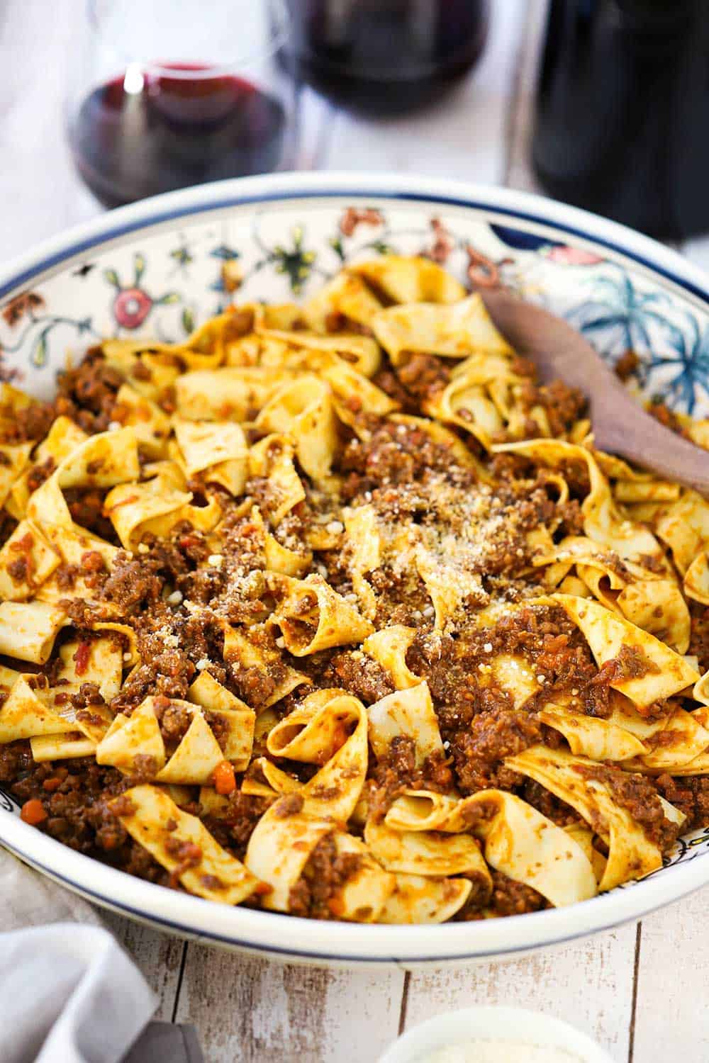 A view looking downward into an Italian pasta bowl filled with pappardelle bolognese pasta next to a couple stemless wine glasses filled with red wine. 