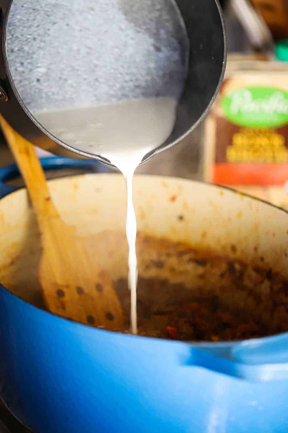 A grey saucepan with warmed milk being poured from it into a blue Dutch oven filled with an Italian meat sauce. 