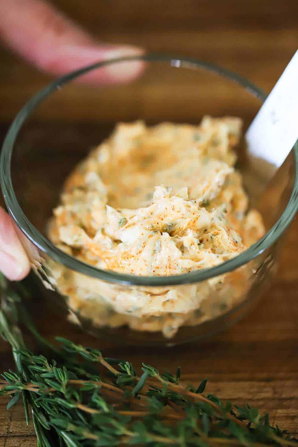 A small glass bowl filled with softened butter that has been blended with fresh herbs, minced garlic, and smoked paprika, all sitting next to a sprig of thyme. 