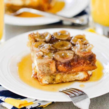 A piece of Caramel Banana Baked French Toast on a white plate with a fork on it and a glass of orange juice in the background.