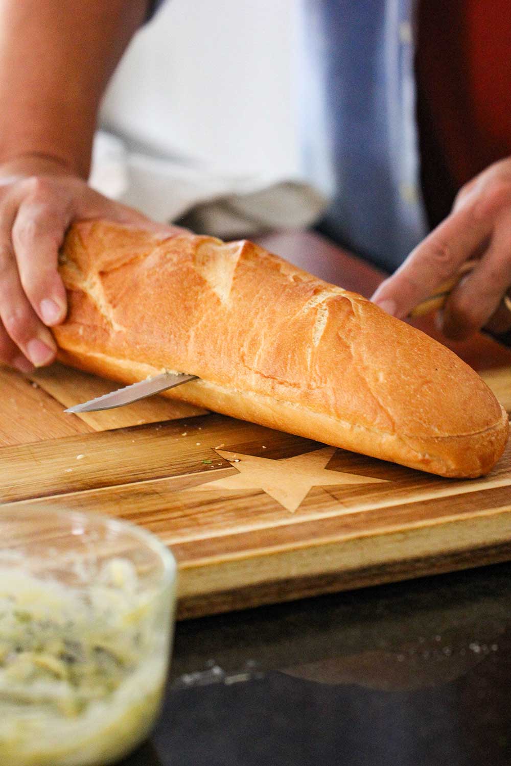 A serrated knife slicing an Italian loaf of bread down the middle on a cutting board. 
