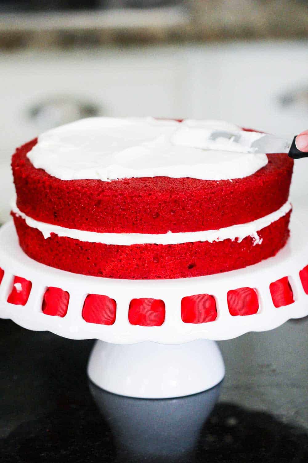 Two layers of red velvet cake being iced while sitting on a cake stand with red ribbon on it. 