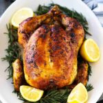 A roast chicken sitting on an oval platter with sliced lemons and herbs surrounding the bird.