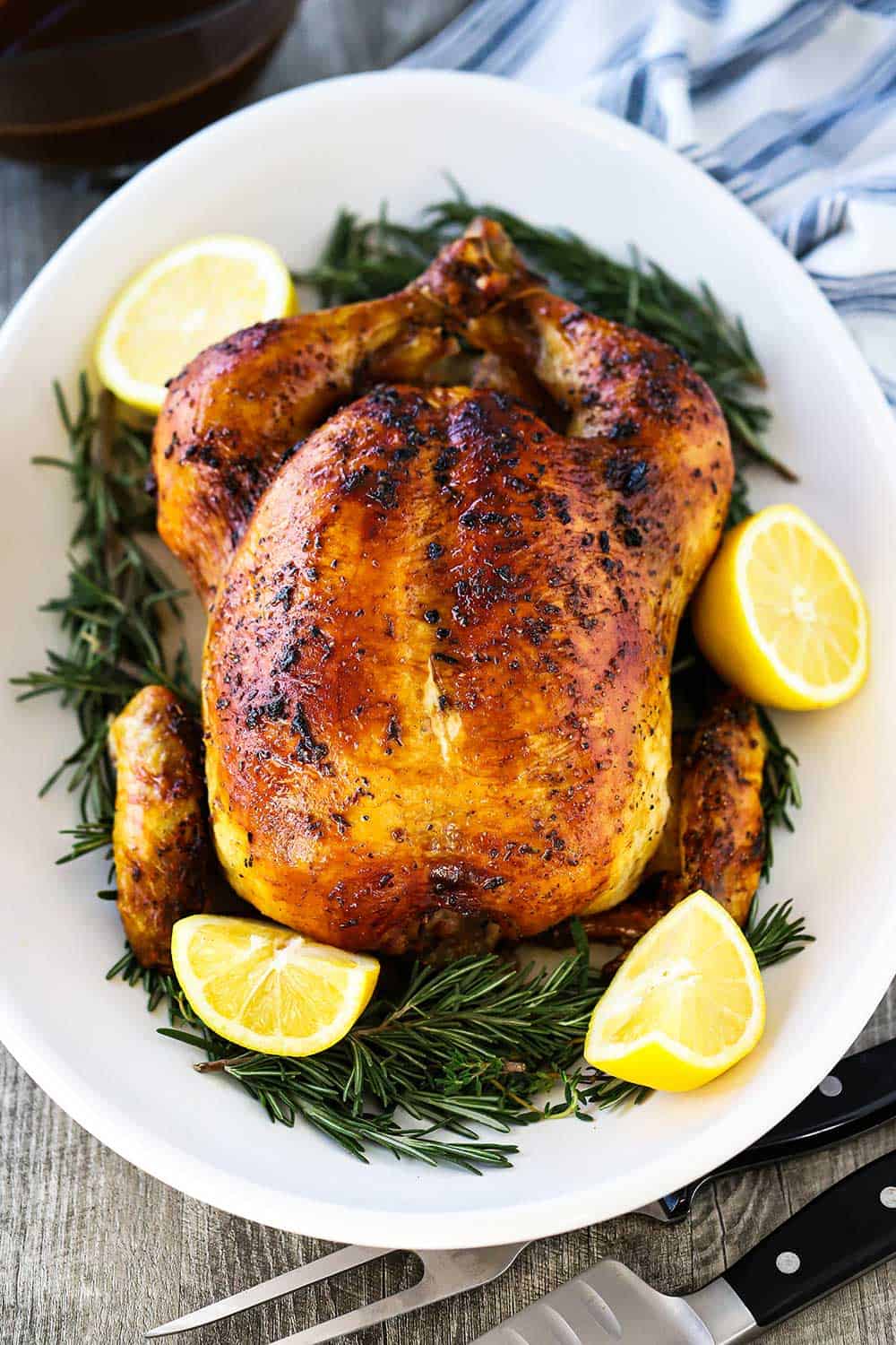 A roast chicken sitting on an oval platter with sliced lemons and herbs surrounding the bird. 