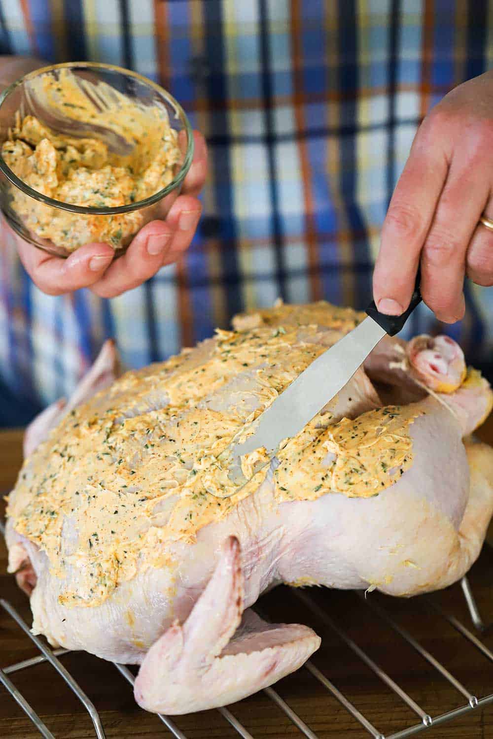 A person using a small metal spatula to spread a herb-butter compound over a whole uncooked turkey sitting on a baking rack. 