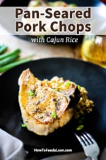 A pan-seared pork chop sitting on a bed of rice on a black dinner plate with a glass of white wine nearby.