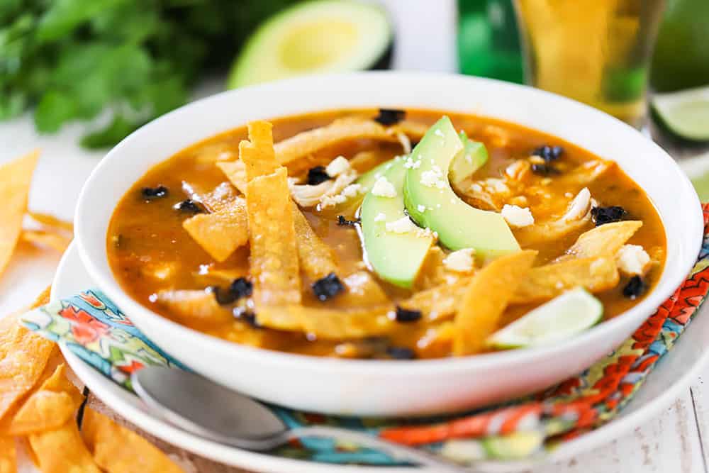 A bowl of chicken tortilla soup in a white bowl surround by a glass of beer and slice avocado.