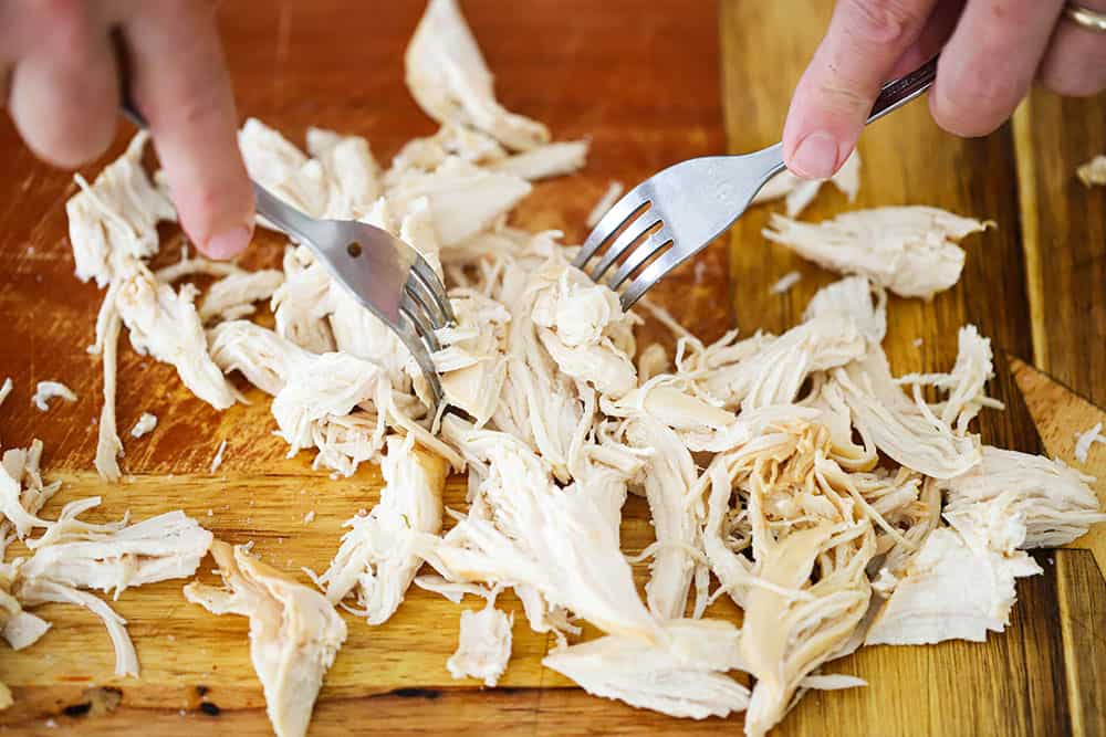 Two forks being used to shred a cooked chicken breast on a cutting board.