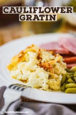 A white dinner plate filled with a serving of cauliflower gratin next to green beans and slices of cooked ham.