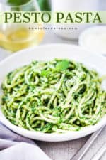 A white bowl filled with pesto pasta next to a grey napkin, two forks, and glass of white wine.