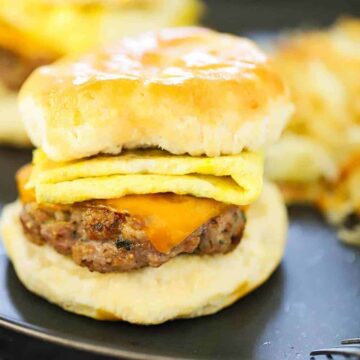 A sausage, egg, and cheese breakfast sandwich on a dark plate in front of hashbrowns.