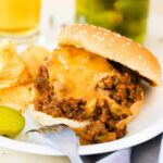 A sloppy joes sandwich on a white plate next to a jar of sliced pickles.
