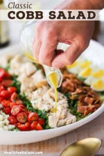 A person pouring a light-colored vinaigrette from a glass salad dressing holder over a platter of a prepared Cobb salad.