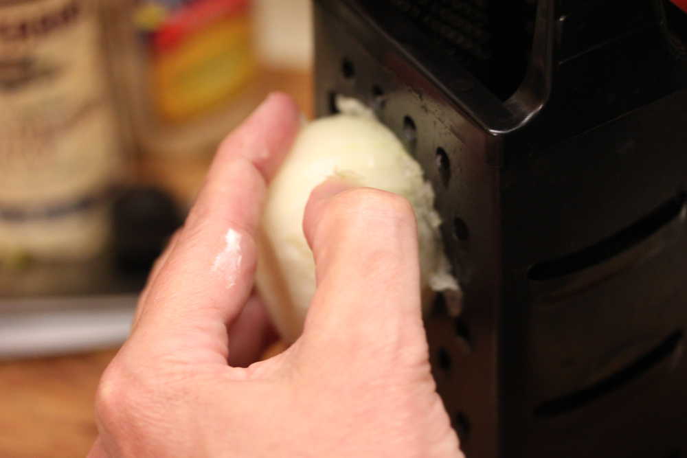 A hand scraping a cut onion down the sides of a box grater, grating the onion. 